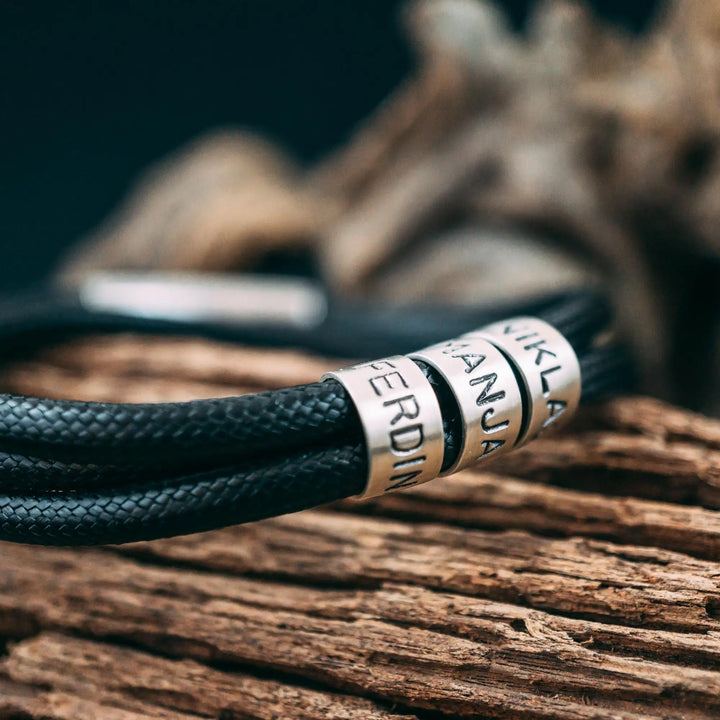 Ein veganes Armband aus schwarzem Synthetik/Baumwoll-Band im Wickelarmband Look, personalisiert mit Gravur von Namen oder Datum auf zylindrischen Sterling Silber 925 Perlen. Das Armband lässt sich mit einem stabilen Verschluss aus Edelstahl verschließen.