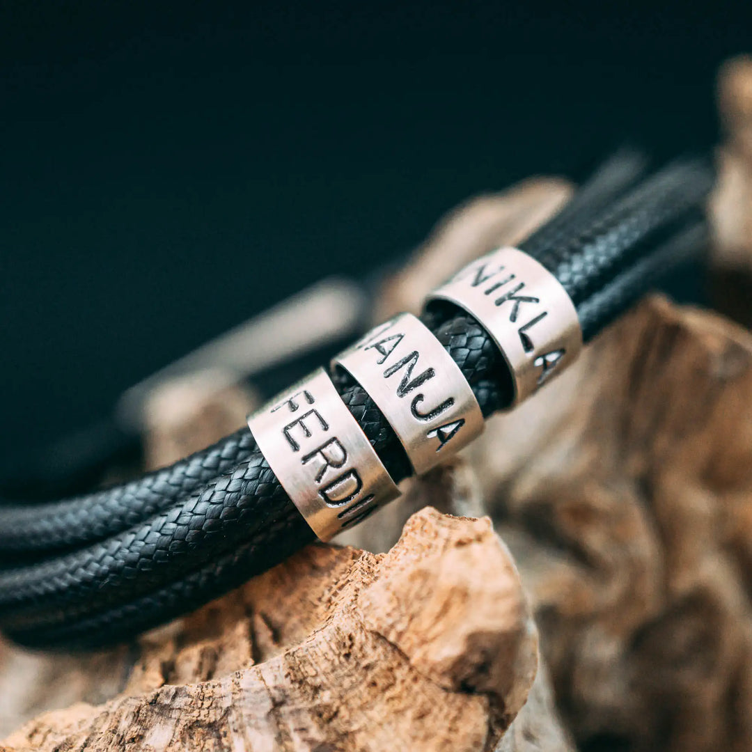Ein veganes Armband aus schwarzem Synthetik/Baumwoll-Band im Wickelarmband Look, personalisiert mit Gravur von Namen oder Datum auf zylindrischen Sterling Silber 925 Perlen. Das Armband lässt sich mit einem stabilen Verschluss aus Edelstahl verschließen.