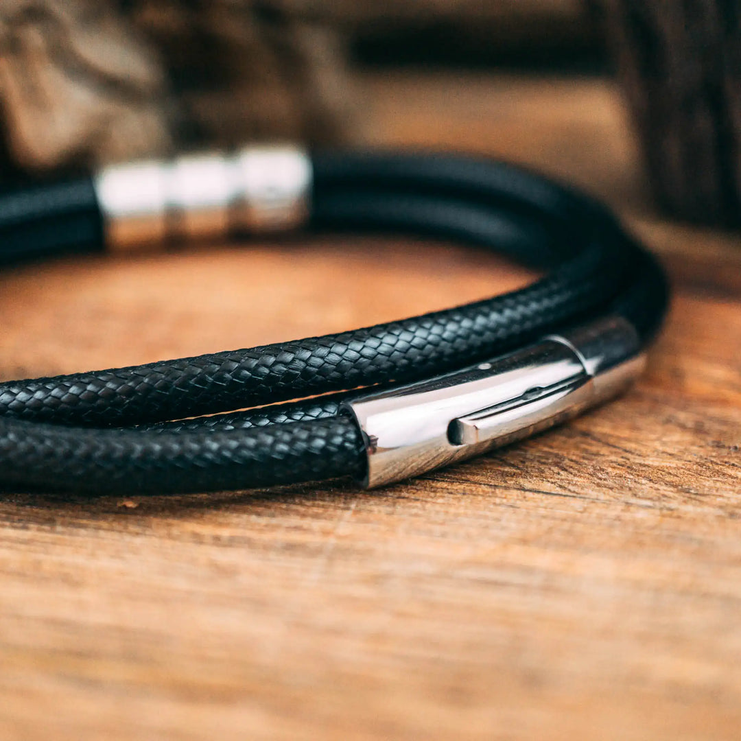 Ein veganes Armband aus schwarzem Synthetik/Baumwoll-Band im Wickelarmband Look, personalisiert mit Gravur von Namen oder Datum auf zylindrischen Sterling Silber 925 Perlen. Das Armband lässt sich mit einem stabilen Verschluss aus Edelstahl verschließen.