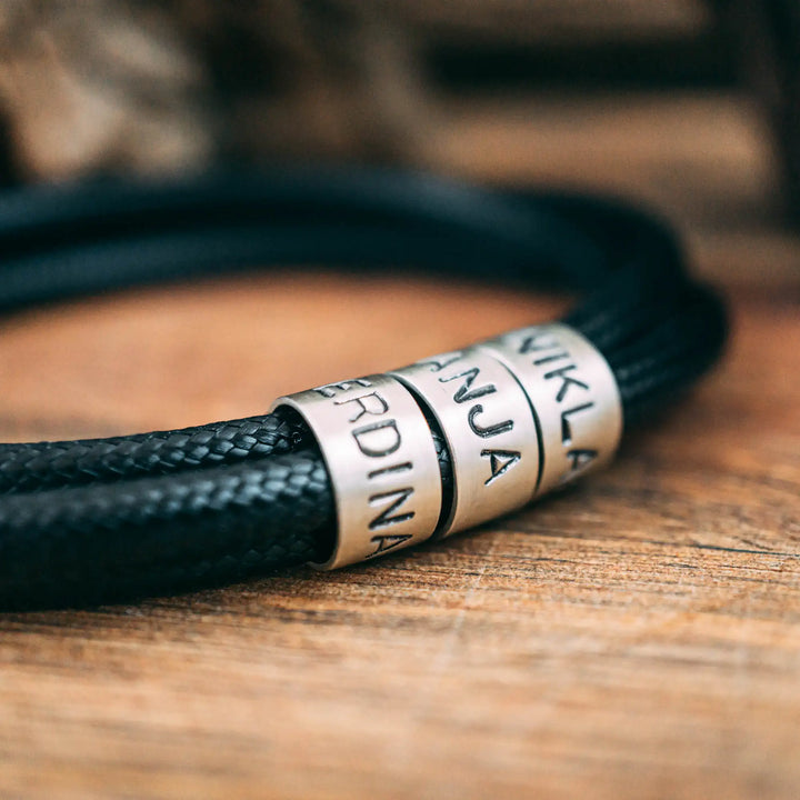 Ein veganes Armband aus schwarzem Synthetik/Baumwoll-Band im Wickelarmband Look, personalisiert mit Gravur von Namen oder Datum auf zylindrischen Sterling Silber 925 Perlen. Das Armband lässt sich mit einem stabilen Verschluss aus Edelstahl verschließen.
