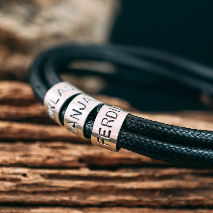 Ein veganes Armband aus schwarzem Synthetik/Baumwoll-Band im Wickelarmband Look, personalisiert mit Gravur von Namen oder Datum auf zylindrischen Sterling Silber 925 Perlen. Das Armband lässt sich mit einem stabilen Verschluss aus Edelstahl verschließen.