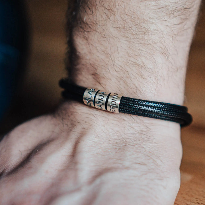 Ein veganes Armband aus schwarzem Synthetik/Baumwoll-Band im Wickelarmband Look, personalisiert mit Gravur von Namen oder Datum auf zylindrischen Sterling Silber 925 Perlen. Das Armband lässt sich mit einem stabilen Verschluss aus Edelstahl verschließen.