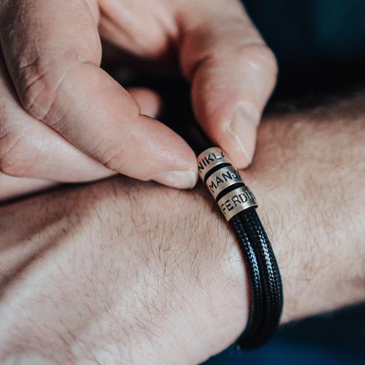 Ein veganes Armband aus schwarzem Synthetik/Baumwoll-Band im Wickelarmband Look, personalisiert mit Gravur von Namen oder Datum auf zylindrischen Sterling Silber 925 Perlen. Das Armband lässt sich mit einem stabilen Verschluss aus Edelstahl verschließen.