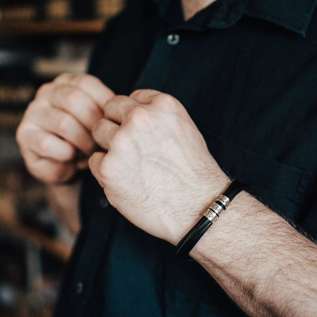 Ein veganes Armband aus schwarzem Synthetik/Baumwoll-Band im Wickelarmband Look, personalisiert mit Gravur von Namen oder Datum auf zylindrischen Sterling Silber 925 Perlen. Das Armband lässt sich mit einem stabilen Verschluss aus Edelstahl verschließen.