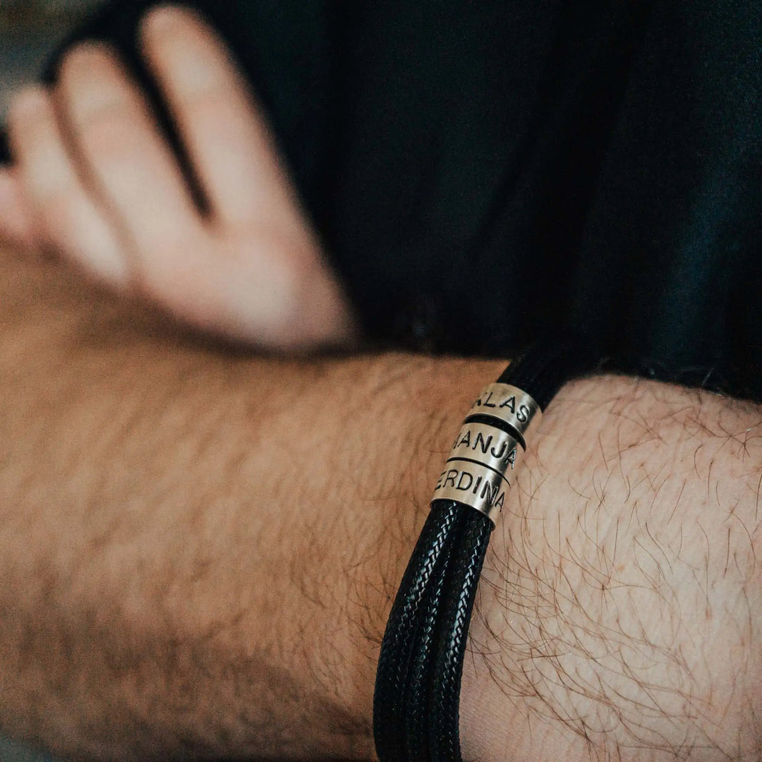 Ein veganes Armband aus schwarzem Synthetik/Baumwoll-Band im Wickelarmband Look, personalisiert mit Gravur von Namen oder Datum auf zylindrischen Sterling Silber 925 Perlen. Das Armband lässt sich mit einem stabilen Verschluss aus Edelstahl verschließen.
