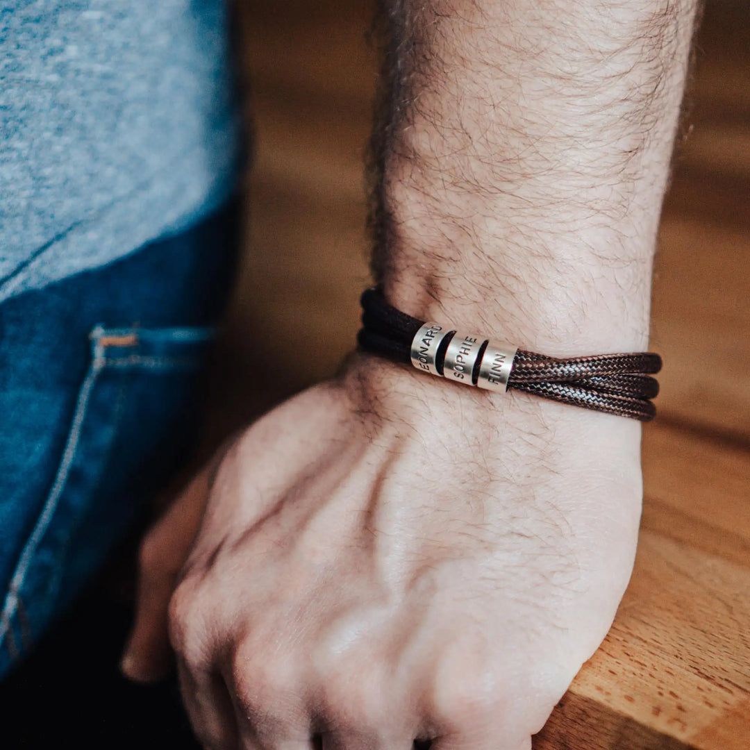 Ein veganes Armband aus braunem Synthetik/Baumwoll-Band im Wickelarmband Look, personalisiert mit Gravur von Namen oder Datum auf runden Sterling Silber 925 Perlen. Das Armband lässt sich mit einem stabilen Verschluss aus Edelstahl verschließen.