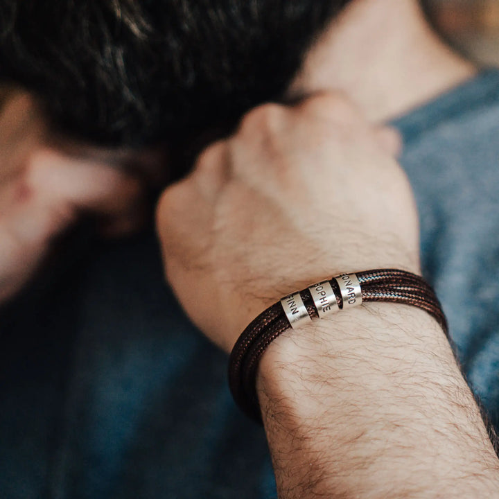 Ein veganes Armband aus braunem Synthetik/Baumwoll-Band im Wickelarmband Look, personalisiert mit Gravur von Namen oder Datum auf runden Sterling Silber 925 Perlen. Das Armband lässt sich mit einem stabilen Verschluss aus Edelstahl verschließen.