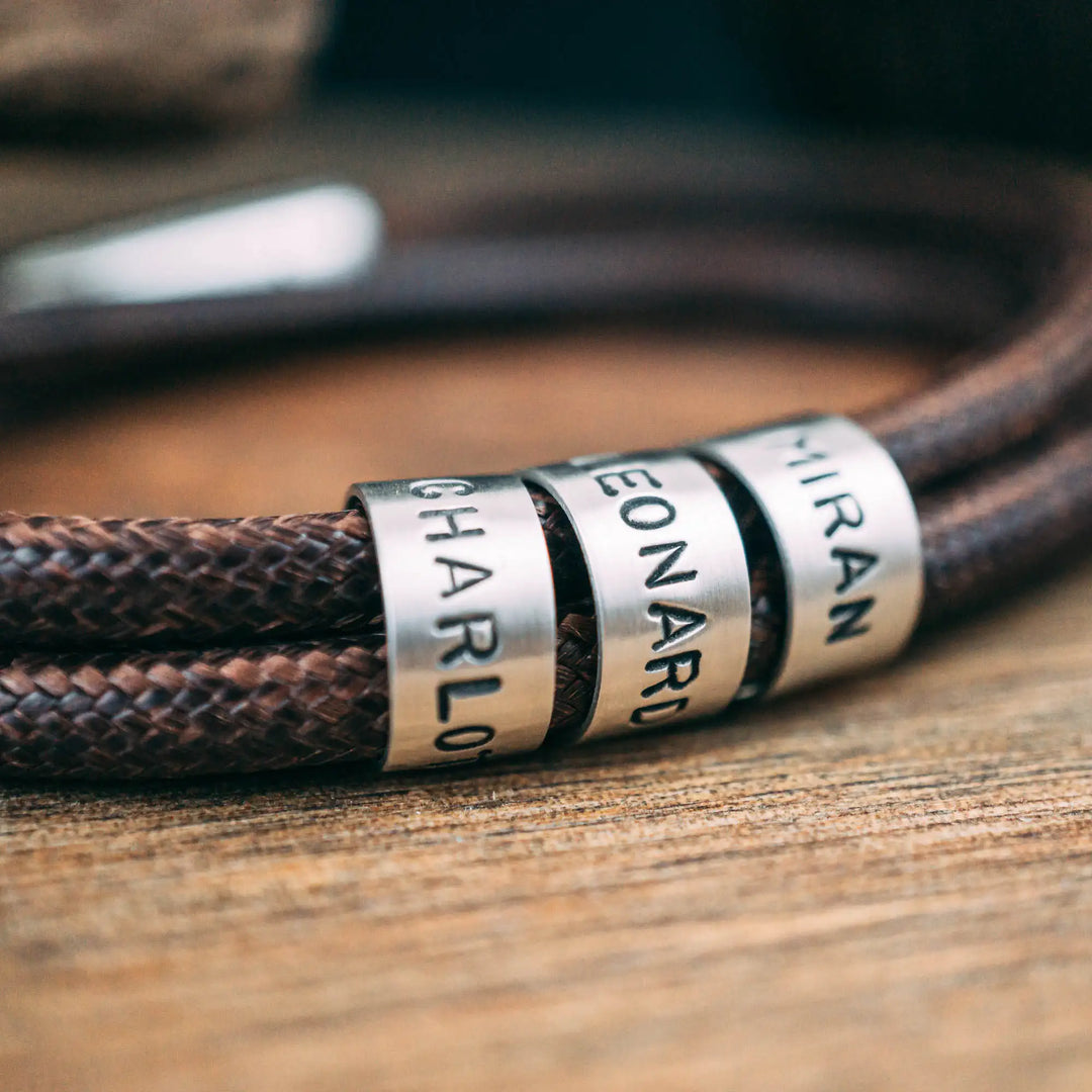 Ein veganes Armband aus braunem Synthetik/Baumwoll-Band im Wickelarmband Look, personalisiert mit Gravur von Namen oder Datum auf runden Sterling Silber 925 Perlen. Das Armband lässt sich mit einem stabilen Verschluss aus Edelstahl verschließen.