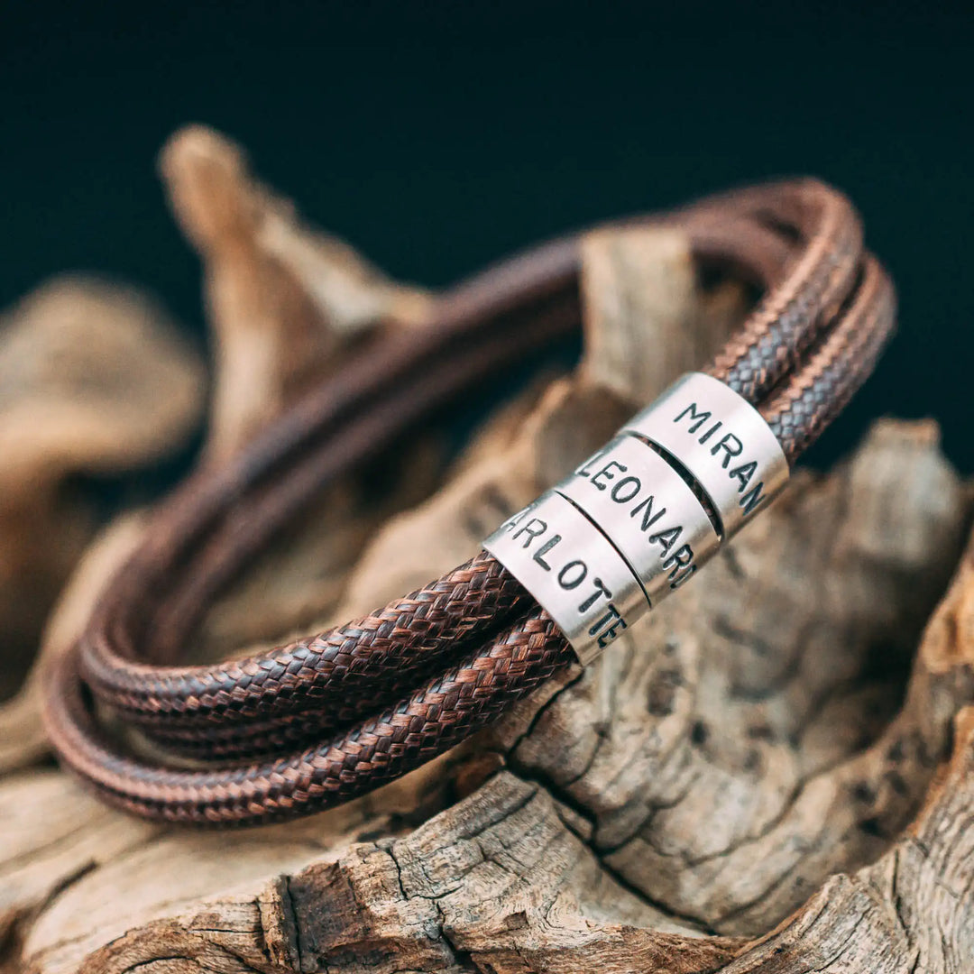 Ein veganes Armband aus braunem Synthetik/Baumwoll-Band im Wickelarmband Look, personalisiert mit Gravur von Namen oder Datum auf runden Sterling Silber 925 Perlen. Das Armband lässt sich mit einem stabilen Verschluss aus Edelstahl verschließen.