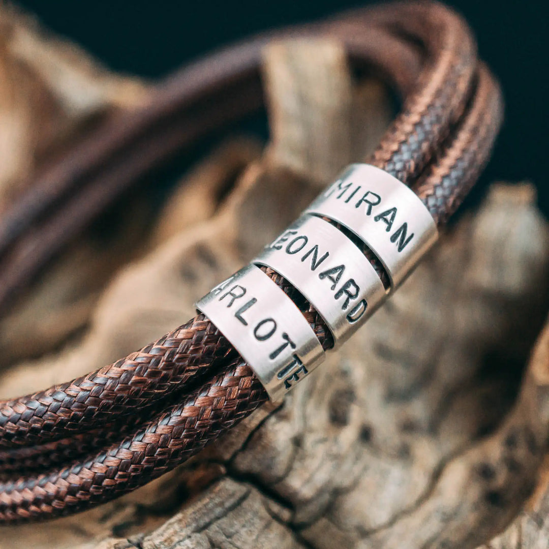 Ein veganes Armband aus braunem Synthetik/Baumwoll-Band im Wickelarmband Look, personalisiert mit Gravur von Namen oder Datum auf runden Sterling Silber 925 Perlen. Das Armband lässt sich mit einem stabilen Verschluss aus Edelstahl verschließen.