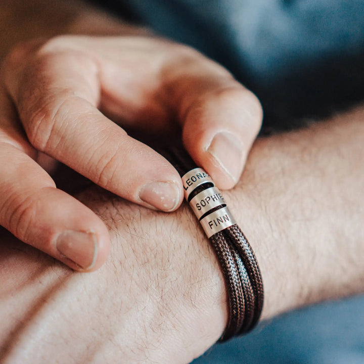 Ein veganes Armband aus braunem Synthetik/Baumwoll-Band im Wickelarmband Look, personalisiert mit Gravur von Namen oder Datum auf runden Sterling Silber 925 Perlen. Das Armband lässt sich mit einem stabilen Verschluss aus Edelstahl verschließen.