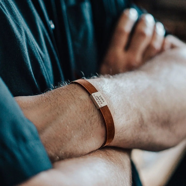 Ein schmales, flaches Armband für Herren aus braunem Leder, personalisiert mit Gravur von Namen oder Datum auf einer rechteckigen Sterling Silber 925 Perle. Das Armband lässt sich mit einem stabilen Verschluss aus Edelstahl verschließen und selbst auf die gewünschte Länge kürzen, was es zum perfekten Geschenk macht.
