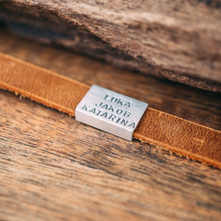 Ein schmales, flaches Armband für Herren aus braunem Leder, personalisiert mit Gravur von Namen oder Datum auf einer rechteckigen Sterling Silber 925 Perle. Das Armband lässt sich mit einem stabilen Verschluss aus Edelstahl verschließen und selbst auf die gewünschte Länge kürzen, was es zum perfekten Geschenk macht.