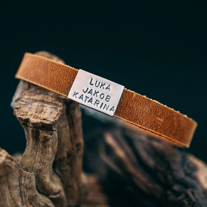 Ein schmales, flaches Armband für Herren aus braunem Leder, personalisiert mit Gravur von Namen oder Datum auf einer rechteckigen Sterling Silber 925 Perle. Das Armband lässt sich mit einem stabilen Verschluss aus Edelstahl verschließen und selbst auf die gewünschte Länge kürzen, was es zum perfekten Geschenk macht.