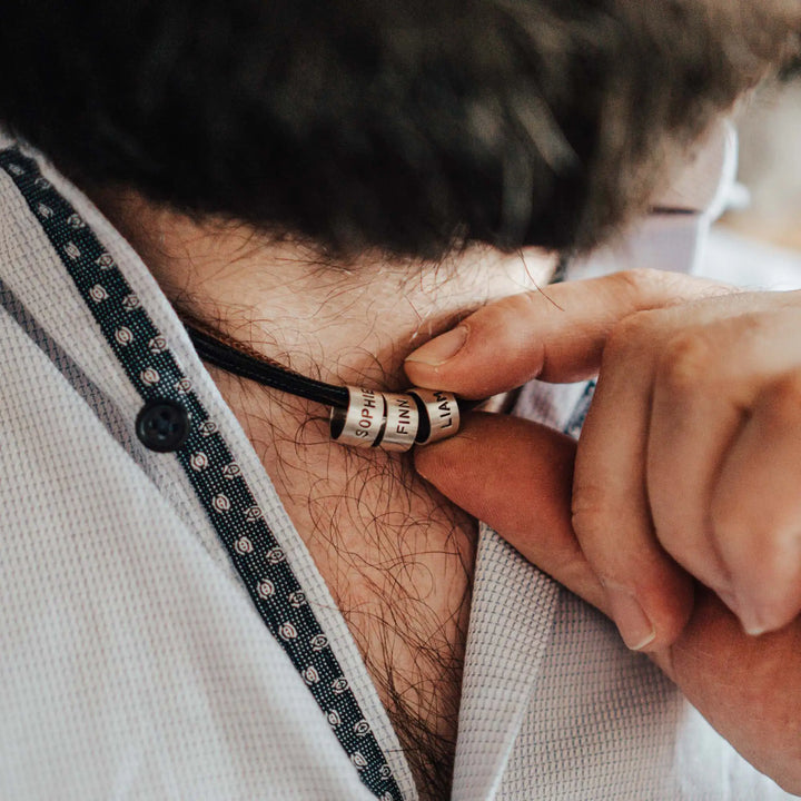 Eine vegane Halskette für Männer aus mehreren braunen und schwarzen Synthetik/Baumwoll-Bändern. Die Kette für Herren ist personalisiert mit Gravur von Namen oder Datum auf zylindrischen Sterling Silber 925 Perlen. Das Kette lässt sich mit einem stabilen Karabiner Verschluss aus Edelstahl verschließen.