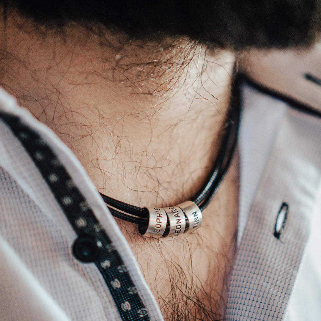 Eine vegane Halskette für Männer aus mehreren schwarzen Synthetik/Baumwoll-Bändern. Die Kette für Herren ist personalisiert mit Gravur von Namen oder Datum auf zylindrischen Sterling Silber 925 Perlen. Das Kette lässt sich mit einem stabilen Karabiner Verschluss aus Edelstahl verschließen.