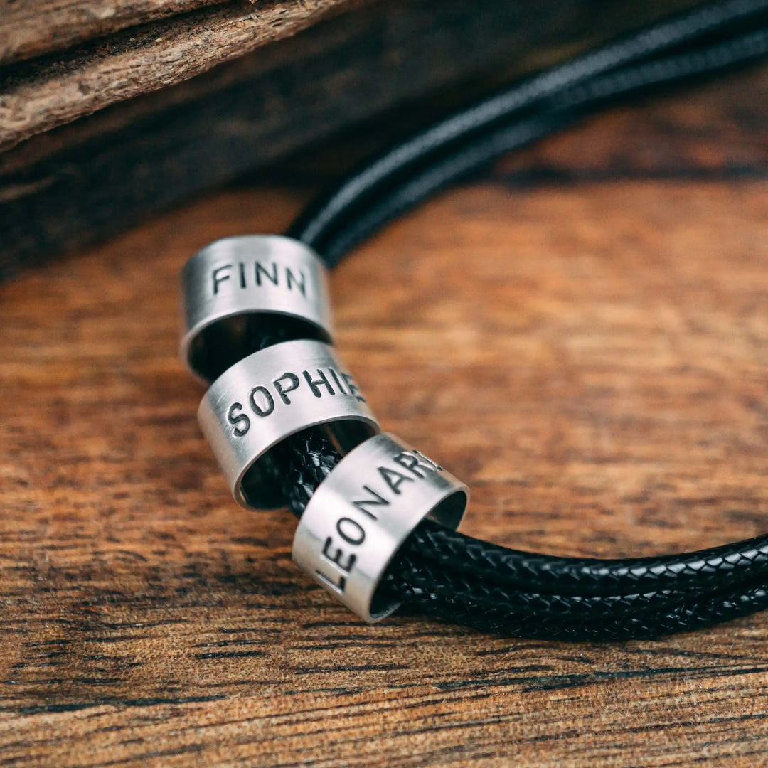 Eine vegane Halskette für Männer aus mehreren schwarzen Synthetik/Baumwoll-Bändern. Die Kette für Herren ist personalisiert mit Gravur von Namen oder Datum auf zylindrischen Sterling Silber 925 Perlen. Das Kette lässt sich mit einem stabilen Karabiner Verschluss aus Edelstahl verschließen.
