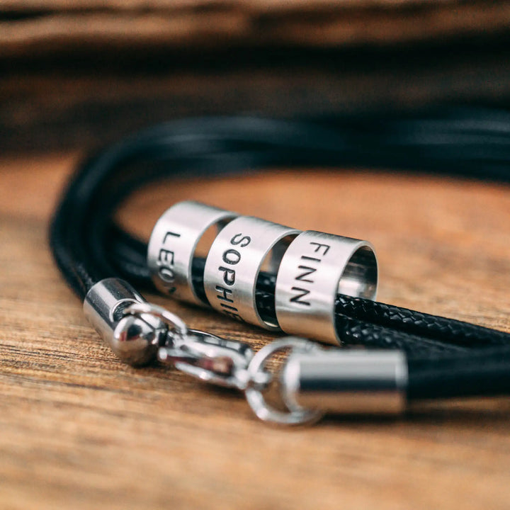 Eine vegane Halskette für Männer aus mehreren schwarzen Synthetik/Baumwoll-Bändern. Die Kette für Herren ist personalisiert mit Gravur von Namen oder Datum auf zylindrischen Sterling Silber 925 Perlen. Das Kette lässt sich mit einem stabilen Karabiner Verschluss aus Edelstahl verschließen.