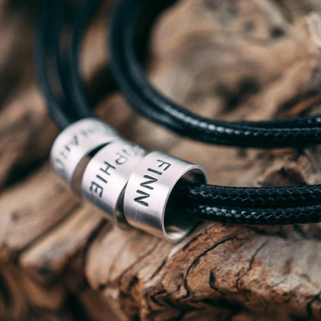 Eine vegane Halskette für Männer aus mehreren schwarzen Synthetik/Baumwoll-Bändern. Die Kette für Herren ist personalisiert mit Gravur von Namen oder Datum auf zylindrischen Sterling Silber 925 Perlen. Das Kette lässt sich mit einem stabilen Karabiner Verschluss aus Edelstahl verschließen.