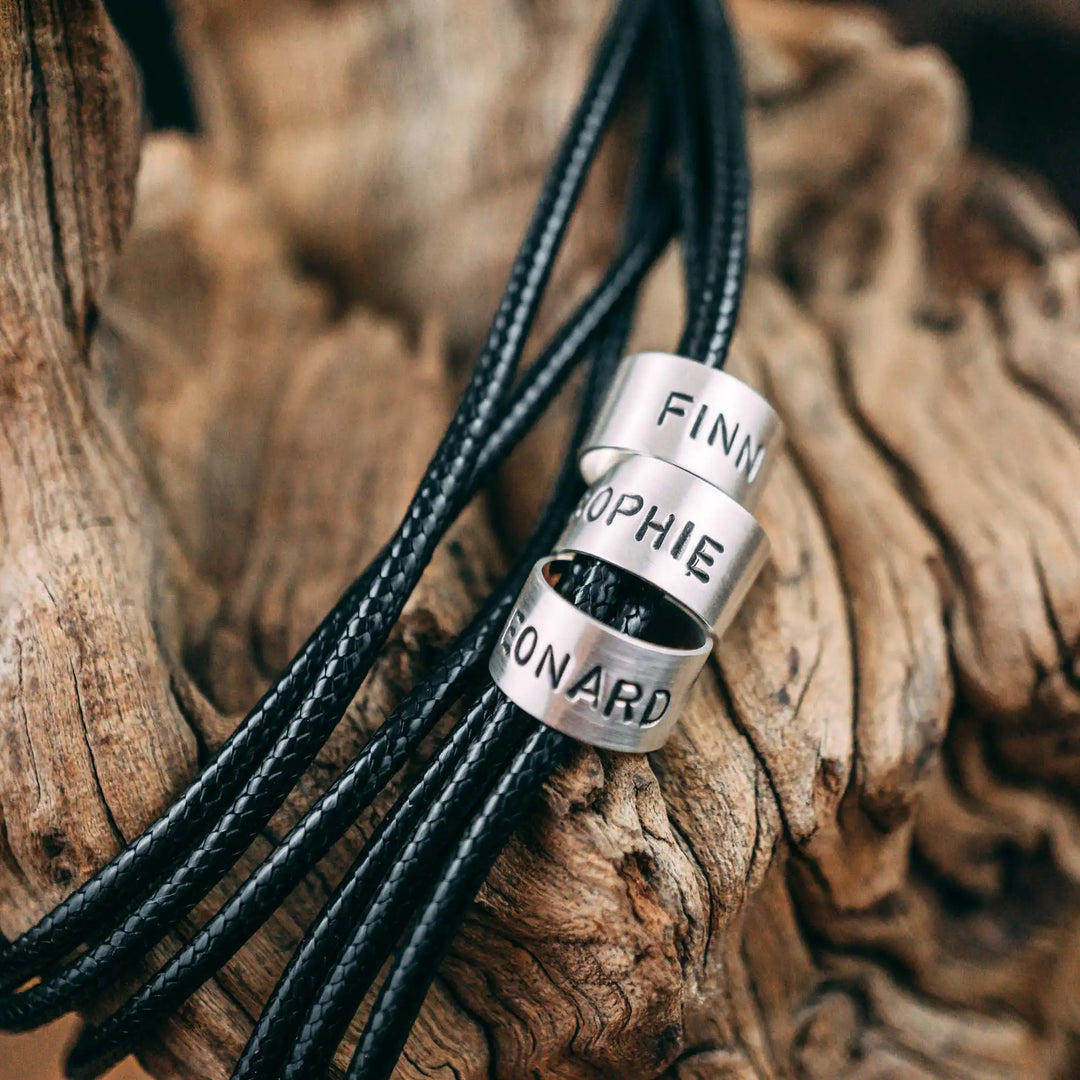 Eine vegane Halskette für Männer aus mehreren schwarzen Synthetik/Baumwoll-Bändern. Die Kette für Herren ist personalisiert mit Gravur von Namen oder Datum auf zylindrischen Sterling Silber 925 Perlen. Das Kette lässt sich mit einem stabilen Karabiner Verschluss aus Edelstahl verschließen.