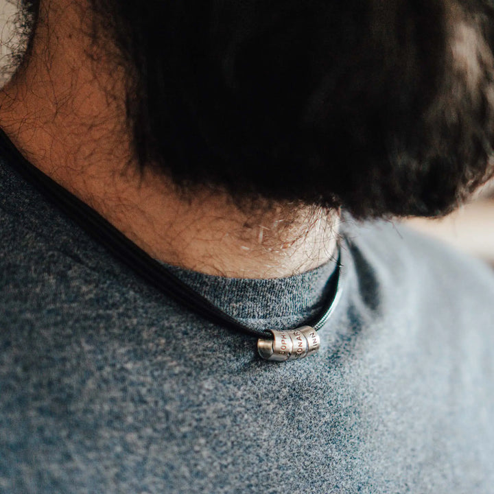 Eine vegane Halskette für Männer aus mehreren schwarzen Synthetik/Baumwoll-Bändern. Die Kette für Herren ist personalisiert mit Gravur von Namen oder Datum auf zylindrischen Sterling Silber 925 Perlen. Das Kette lässt sich mit einem stabilen Karabiner Verschluss aus Edelstahl verschließen.