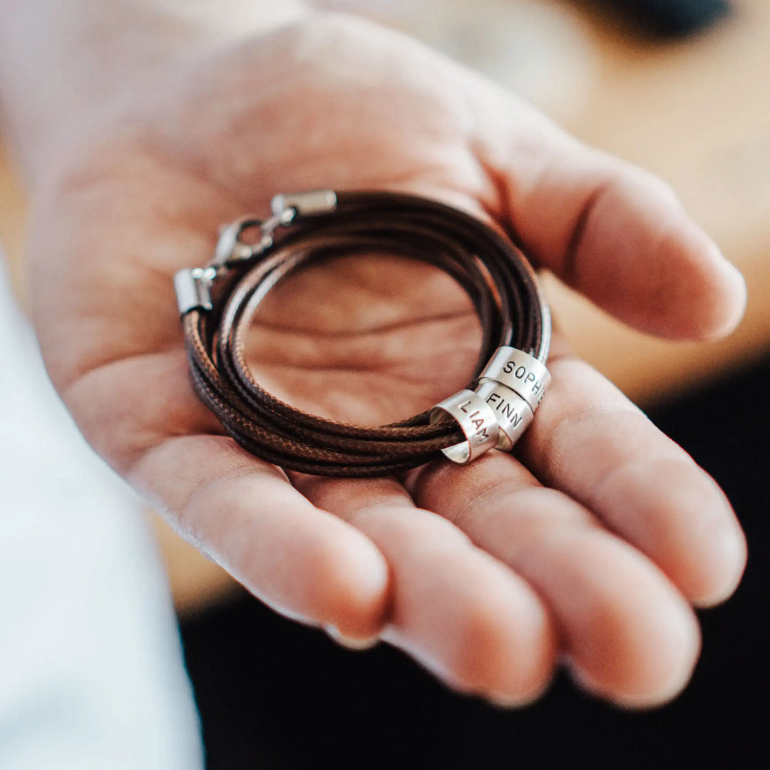 Eine vegane Halskette für Männer aus mehreren braunen Synthetik/Baumwoll-Bändern. Die Kette für Herren ist personalisiert mit Gravur von Namen oder Datum auf zylindrischen Sterling Silber 925 Perlen. Das Kette lässt sich mit einem stabilen Karabiner Verschluss aus Edelstahl verschließen.