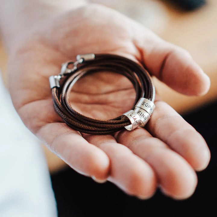 Eine vegane Halskette für Männer aus mehreren braunen Synthetik/Baumwoll-Bändern. Die Kette für Herren ist personalisiert mit Gravur von Namen oder Datum auf zylindrischen Sterling Silber 925 Perlen. Das Kette lässt sich mit einem stabilen Karabiner Verschluss aus Edelstahl verschließen.