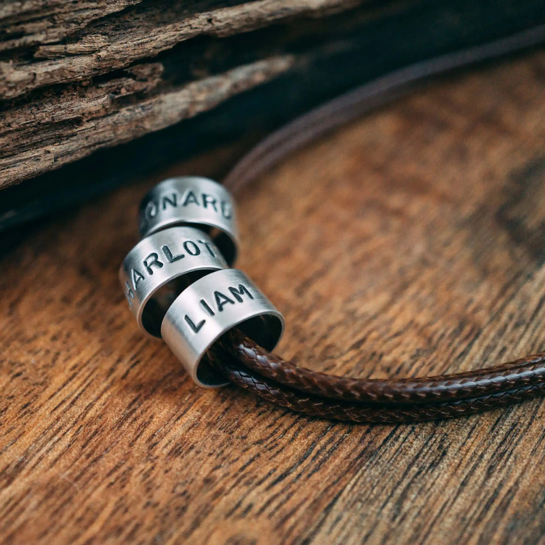 Eine vegane Halskette für Männer aus mehreren braunen Synthetik/Baumwoll-Bändern. Die Kette für Herren ist personalisiert mit Gravur von Namen oder Datum auf zylindrischen Sterling Silber 925 Perlen. Das Kette lässt sich mit einem stabilen Karabiner Verschluss aus Edelstahl verschließen.