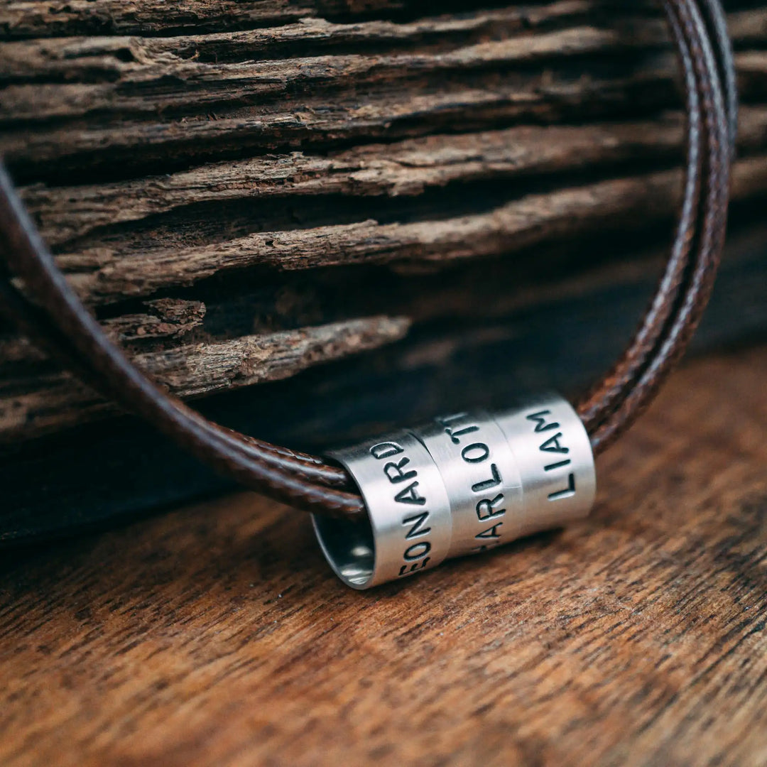 Eine vegane Halskette für Männer aus mehreren braunen Synthetik/Baumwoll-Bändern. Die Kette für Herren ist personalisiert mit Gravur von Namen oder Datum auf zylindrischen Sterling Silber 925 Perlen. Das Kette lässt sich mit einem stabilen Karabiner Verschluss aus Edelstahl verschließen.