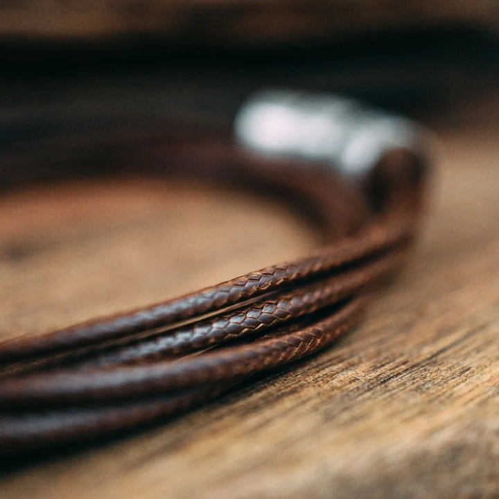 Eine vegane Halskette für Männer aus mehreren braunen Synthetik/Baumwoll-Bändern. Die Kette für Herren ist personalisiert mit Gravur von Namen oder Datum auf zylindrischen Sterling Silber 925 Perlen. Das Kette lässt sich mit einem stabilen Karabiner Verschluss aus Edelstahl verschließen.