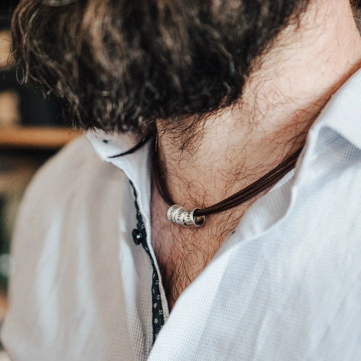 Eine vegane Halskette für Männer aus mehreren braunen Synthetik/Baumwoll-Bändern. Die Kette für Herren ist personalisiert mit Gravur von Namen oder Datum auf zylindrischen Sterling Silber 925 Perlen. Das Kette lässt sich mit einem stabilen Karabiner Verschluss aus Edelstahl verschließen.