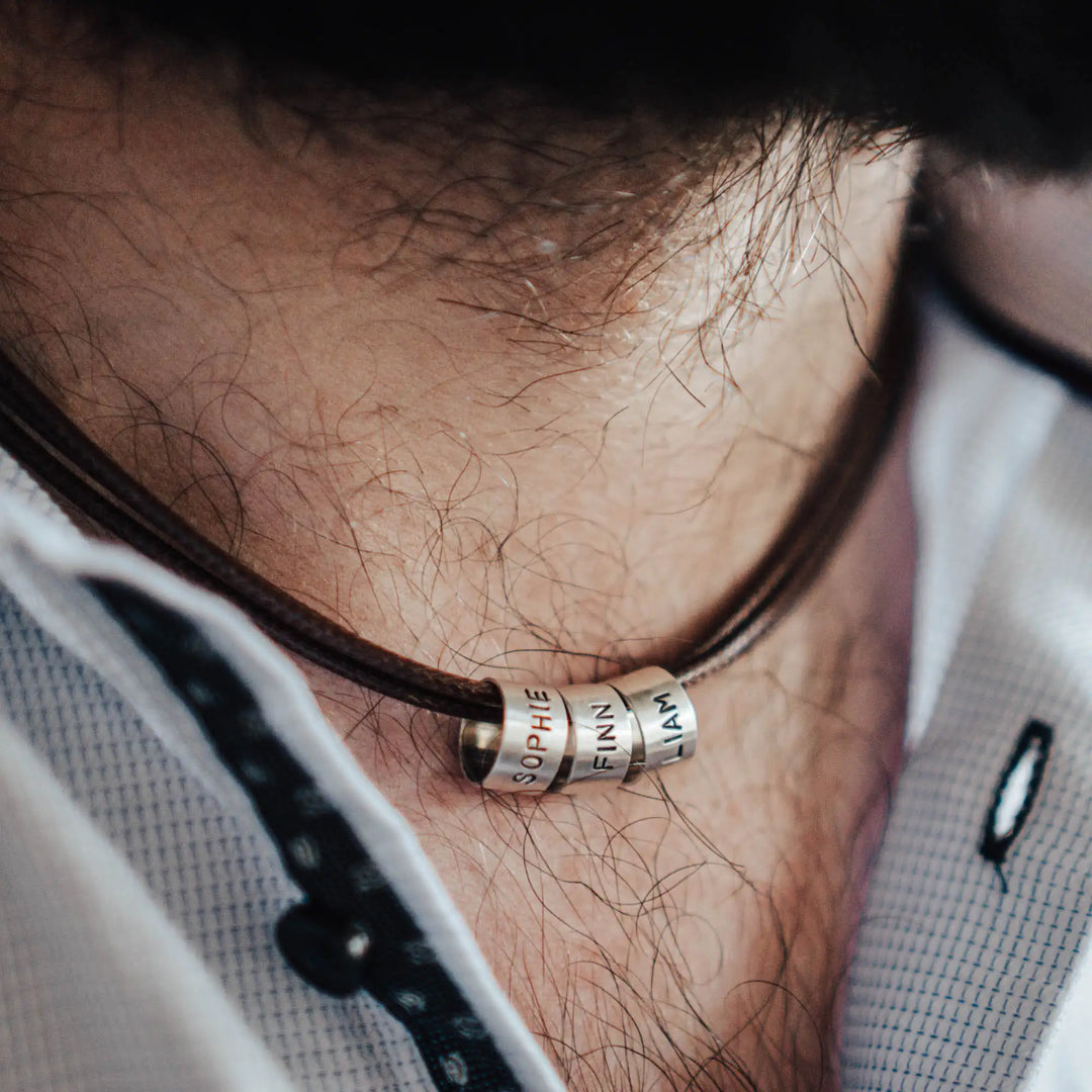 Eine vegane Halskette für Männer aus mehreren braunen Synthetik/Baumwoll-Bändern. Die Kette für Herren ist personalisiert mit Gravur von Namen oder Datum auf zylindrischen Sterling Silber 925 Perlen. Das Kette lässt sich mit einem stabilen Karabiner Verschluss aus Edelstahl verschließen.