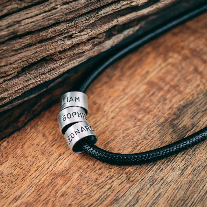 Eine vegane Halskette für Männer aus schwarzem Synthetik/Baumwoll-Band. Die Kette für Herren ist personalisiert mit Gravur von Namen oder Datum auf zylindrischen Sterling Silber 925 Perlen. Das Kette lässt sich mit einem stabilen Karabiner Verschluss aus Edelstahl verschließen.