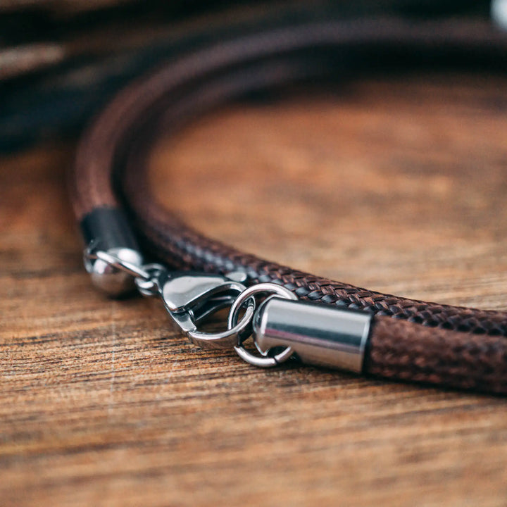 Eine vegane Halskette für Männer aus braunem Synthetik/Baumwoll-Band. Die Kette für Herren ist personalisiert mit Gravur von Namen oder Datum auf zylindrischen Sterling Silber 925 Perlen. Das Kette lässt sich mit einem stabilen Karabiner Verschluss aus Edelstahl verschließen.