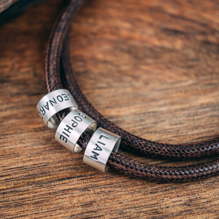 Eine vegane Halskette für Männer aus braunem Synthetik/Baumwoll-Band. Die Kette für Herren ist personalisiert mit Gravur von Namen oder Datum auf zylindrischen Sterling Silber 925 Perlen. Das Kette lässt sich mit einem stabilen Karabiner Verschluss aus Edelstahl verschließen.