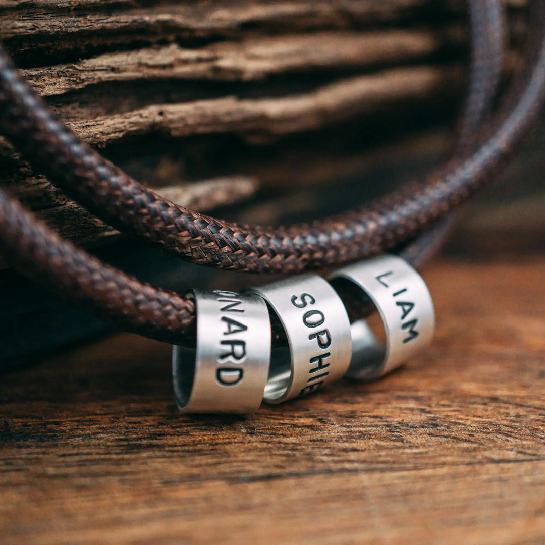 Eine vegane Halskette für Männer aus braunem Synthetik/Baumwoll-Band. Die Kette für Herren ist personalisiert mit Gravur von Namen oder Datum auf zylindrischen Sterling Silber 925 Perlen. Das Kette lässt sich mit einem stabilen Karabiner Verschluss aus Edelstahl verschließen.