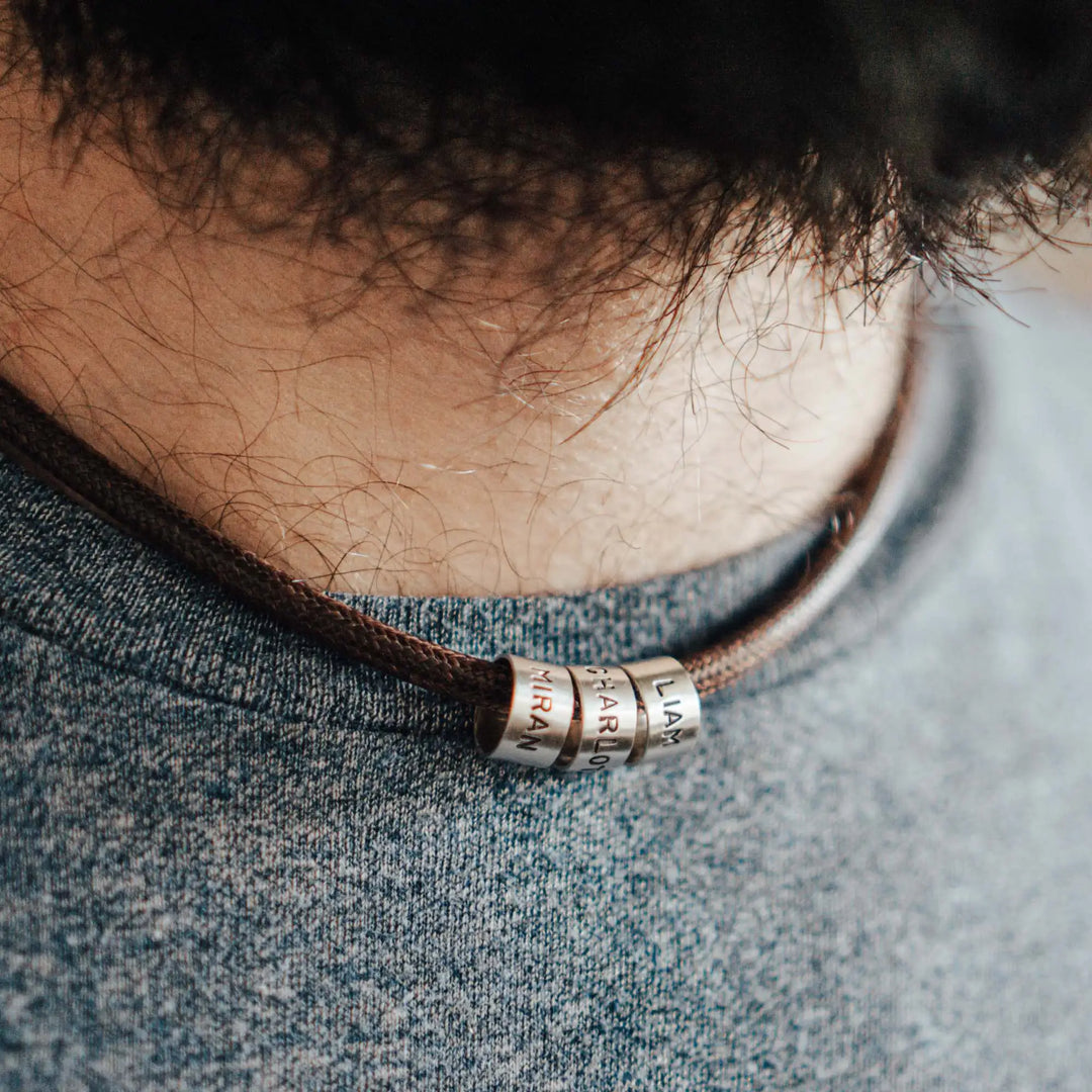 Eine vegane Halskette für Männer aus braunem Synthetik/Baumwoll-Band. Die Kette für Herren ist personalisiert mit Gravur von Namen oder Datum auf zylindrischen Sterling Silber 925 Perlen. Das Kette lässt sich mit einem stabilen Karabiner Verschluss aus Edelstahl verschließen.