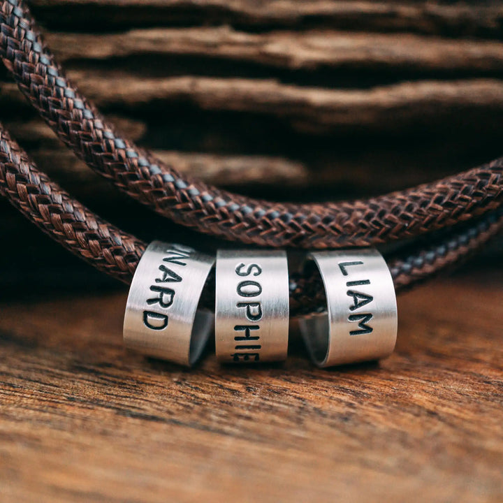 Eine vegane Halskette für Männer aus braunem Synthetik/Baumwoll-Band. Die Kette für Herren ist personalisiert mit Gravur von Namen oder Datum auf zylindrischen Sterling Silber 925 Perlen. Das Kette lässt sich mit einem stabilen Karabiner Verschluss aus Edelstahl verschließen.