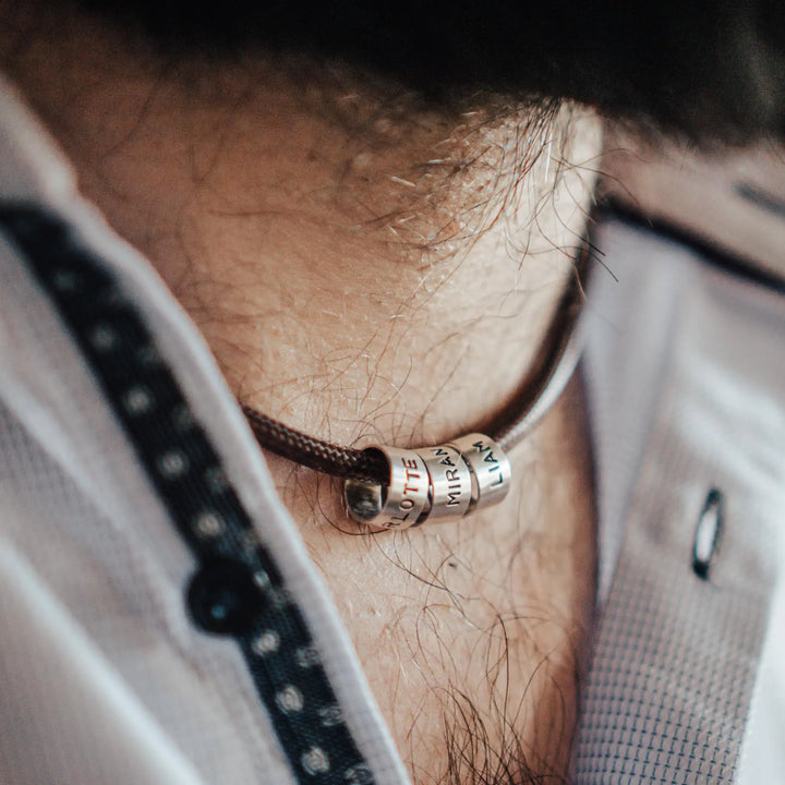 Eine vegane Halskette für Männer aus braunem Synthetik/Baumwoll-Band. Die Kette für Herren ist personalisiert mit Gravur von Namen oder Datum auf zylindrischen Sterling Silber 925 Perlen. Das Kette lässt sich mit einem stabilen Karabiner Verschluss aus Edelstahl verschließen.