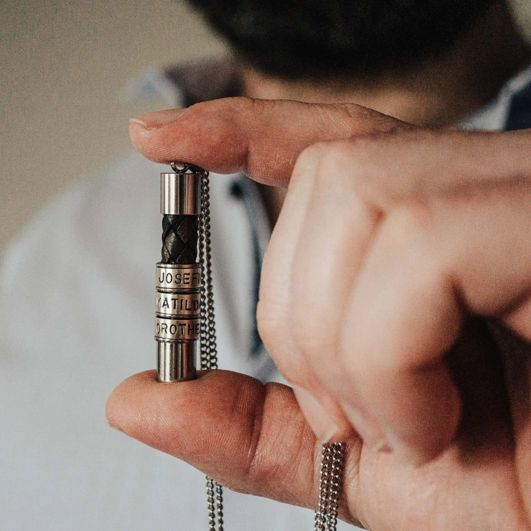 Eine Halskette für Männer aus braunem Leder-Anhänger und einer Edelstahl-Kette. Die Kette für Herren ist an dem zylindrischen Anhänger aus Leder personalisiert mit Gravur von Namen oder Datum auf zylindrischen Sterling Silber 925 Perlen. Die Kette lässt sich mit einem silbernen Karabiner aus Edelstahl verschließen.