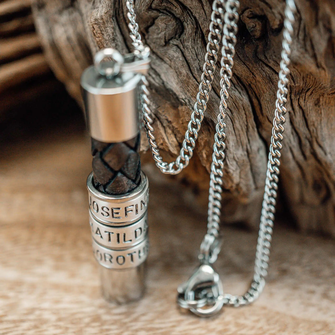 Eine Halskette für Männer aus braunem Leder-Anhänger und einer Edelstahl-Kette. Die Kette für Herren ist an dem zylindrischen Anhänger aus Leder personalisiert mit Gravur von Namen oder Datum auf zylindrischen Sterling Silber 925 Perlen. Die Kette lässt sich mit einem silbernen Karabiner aus Edelstahl verschließen.