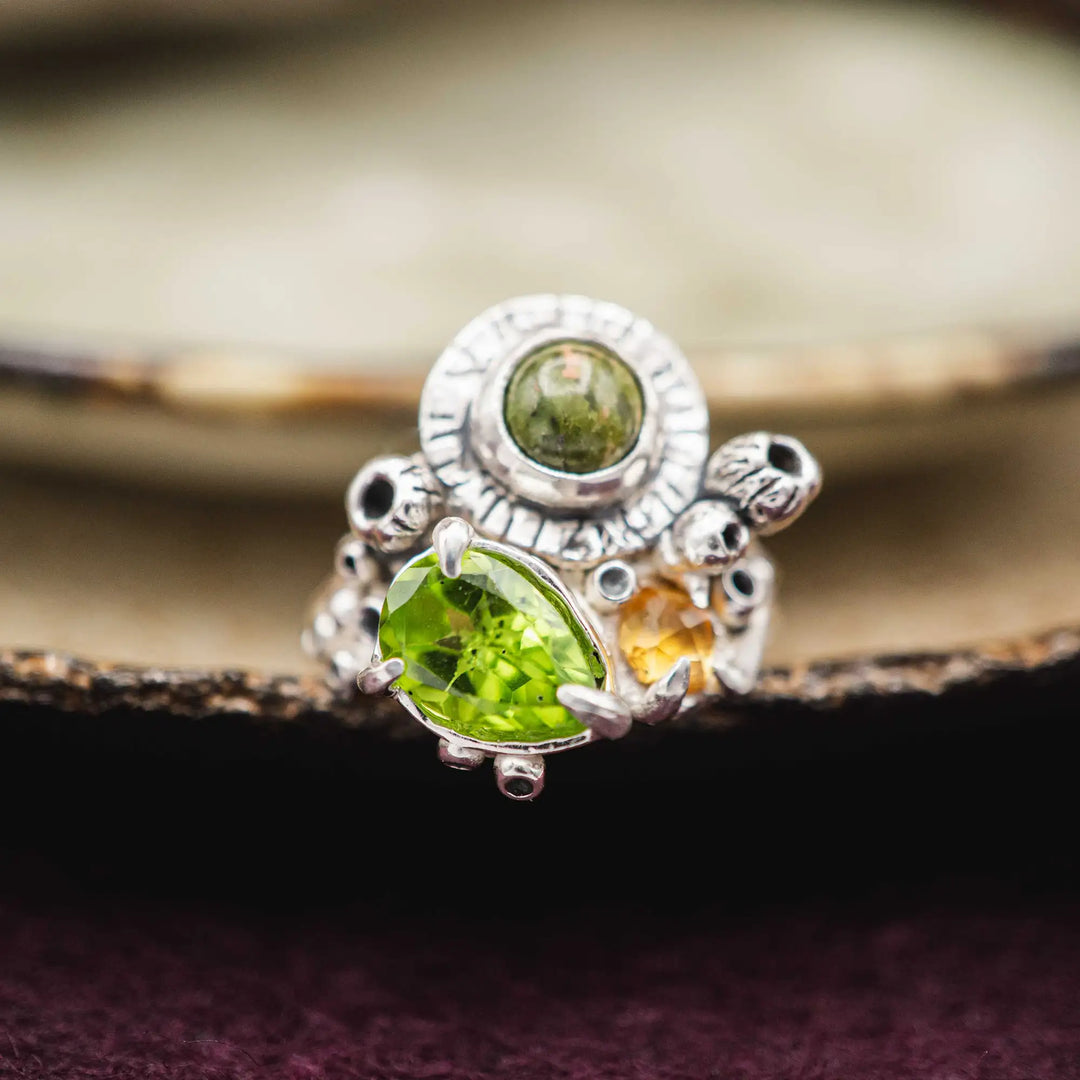 Ring mit 3 Steinen aus Silber. Die Steine sind ein grüner Peridot Tropfen, ein eckiger gelber Citrin und ein runder grüner Unakit. Der Naturschmuck Ring ist rustikal und üppig mit Knospen verziert.