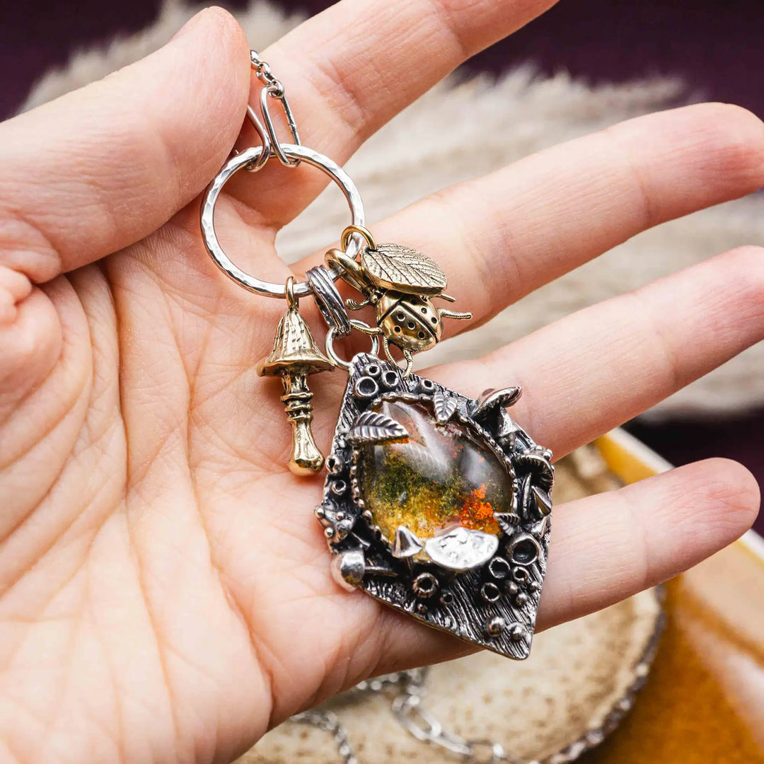 Statement Kette groß aus Silber 925. In den Anhänger eingefasst ist ein tropfenförmiger Lodolith, auch Gartenquarz genannt. Der Stein sieht aus wie eingeschlossenes grünes, gelbes und orangefarbenes Moos und ist umrandet von Pilzen, Blättern und Knospen. Ein echtes, handgefertigtes Statement Piece an Naturschmuck.
An dem Anhänger hängen außerdem 3 kleine Charms aus Messing: Ein Marienkäfer, ein Pilz und ein Blatt.