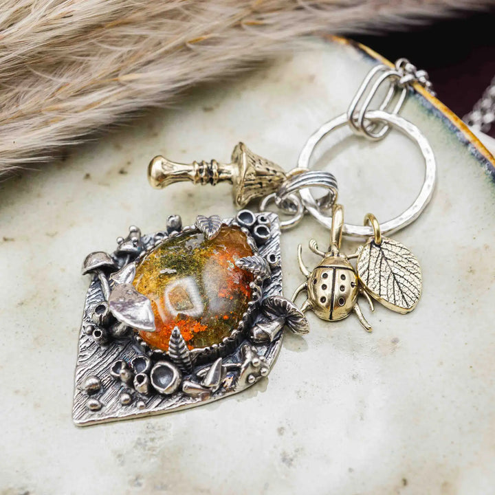 Statement Kette groß aus Silber 925. In den Anhänger eingefasst ist ein tropfenförmiger Lodolith, auch Gartenquarz genannt. Der Stein sieht aus wie eingeschlossenes grünes, gelbes und orangefarbenes Moos und ist umrandet von Pilzen, Blättern und Knospen. Ein echtes, handgefertigtes Statement Piece an Naturschmuck.
An dem Anhänger hängen außerdem 3 kleine Charms aus Messing: Ein Marienkäfer, ein Pilz und ein Blatt.