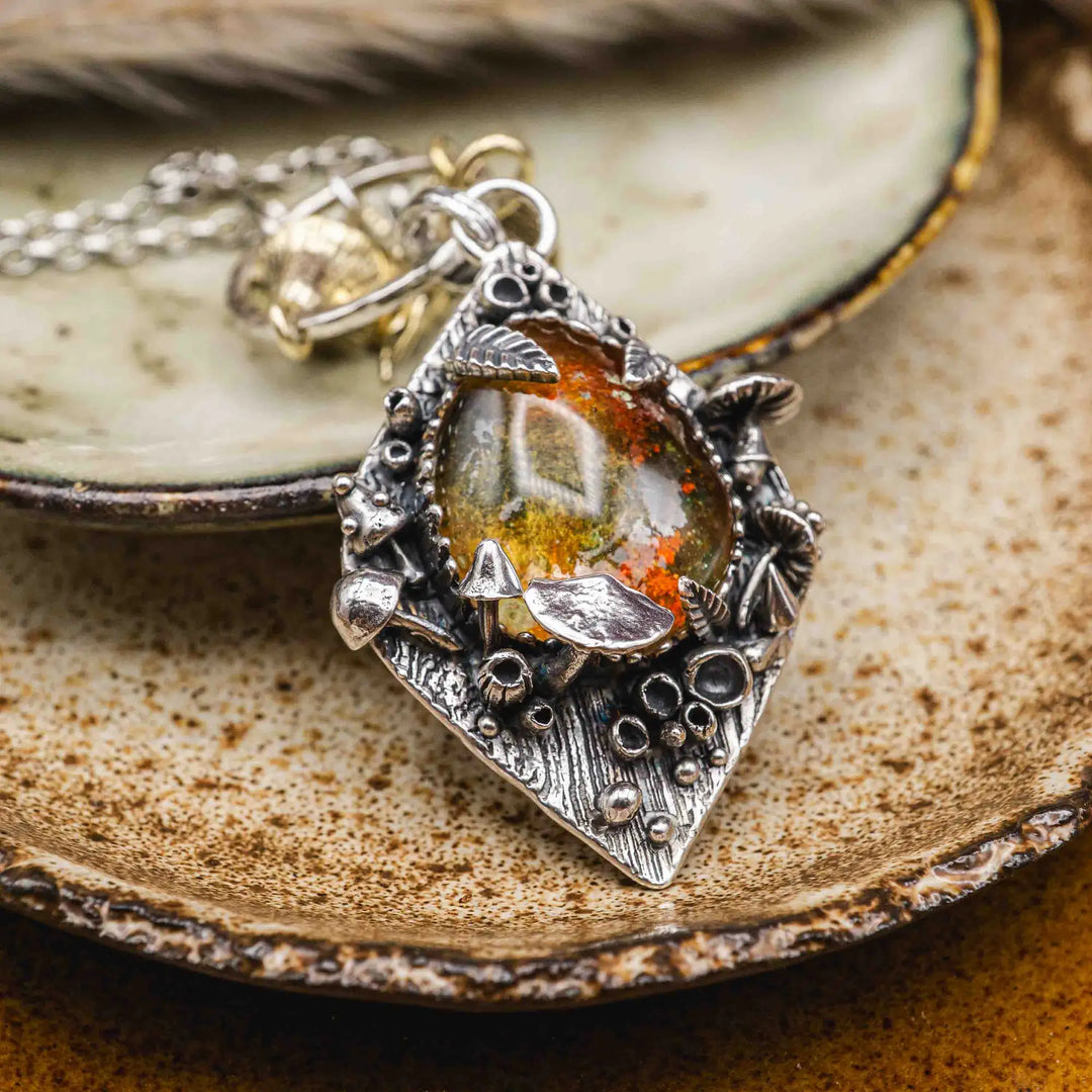 Statement Kette groß aus Silber 925. In den Anhänger eingefasst ist ein tropfenförmiger Lodolith, auch Gartenquarz genannt. Der Stein sieht aus wie eingeschlossenes grünes, gelbes und orangefarbenes Moos und ist umrandet von Pilzen, Blättern und Knospen. Ein echtes, handgefertigtes Statement Piece an Naturschmuck.
An dem Anhänger hängen außerdem 3 kleine Charms aus Messing: Ein Marienkäfer, ein Pilz und ein Blatt.
