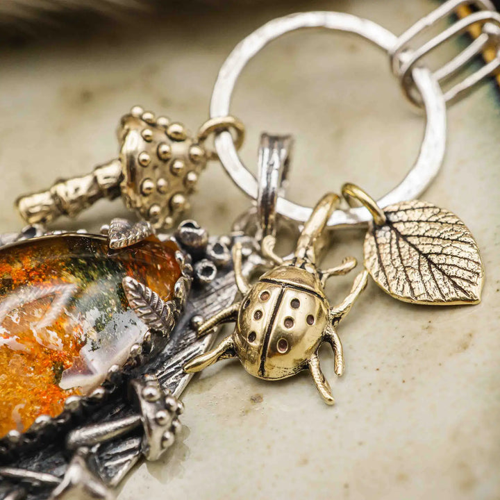 Statement Kette groß aus Silber 925. In den Anhänger eingefasst ist ein tropfenförmiger Lodolith, auch Gartenquarz genannt. Der Stein sieht aus wie eingeschlossenes grünes, gelbes und orangefarbenes Moos und ist umrandet von Pilzen, Blättern und Knospen. Ein echtes, handgefertigtes Statement Piece an Naturschmuck.
An dem Anhänger hängen außerdem 3 kleine Charms aus Messing: Ein Marienkäfer, ein Pilz und ein Blatt.