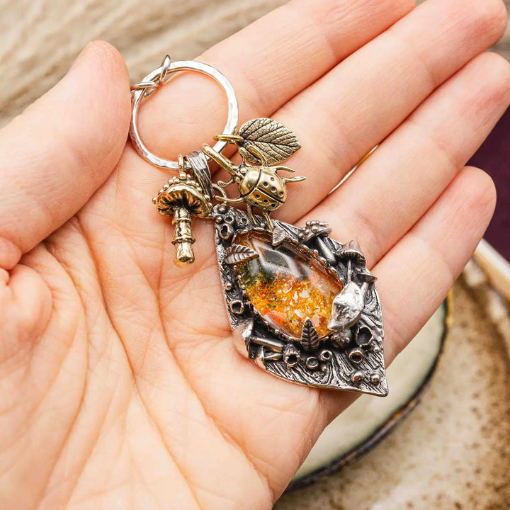 Statement Kette groß aus Silber 925. In den Anhänger eingefasst ist ein tropfenförmiger Lodolith, auch Gartenquarz genannt. Der Stein sieht aus wie eingeschlossenes grünes, gelbes und orangefarbenes Moos und ist umrandet von Pilzen, Blättern und Knospen. Ein echtes, handgefertigtes Statement Piece an Naturschmuck.
An dem Anhänger hängen außerdem 3 kleine Charms aus Messing: Ein Marienkäfer, ein Pilz und ein Blatt.