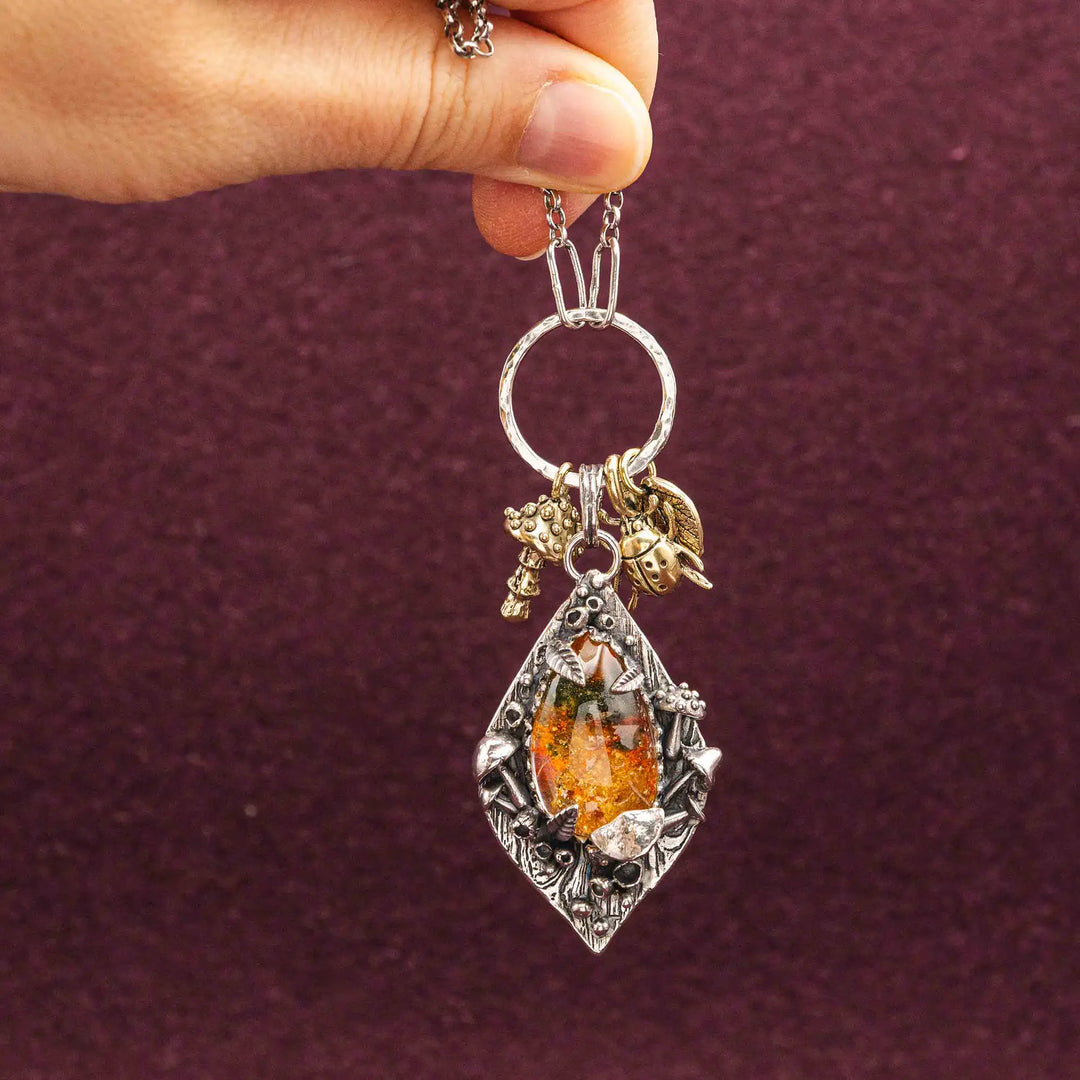 Statement Kette groß aus Silber 925. In den Anhänger eingefasst ist ein tropfenförmiger Lodolith, auch Gartenquarz genannt. Der Stein sieht aus wie eingeschlossenes grünes, gelbes und orangefarbenes Moos und ist umrandet von Pilzen, Blättern und Knospen. Ein echtes, handgefertigtes Statement Piece an Naturschmuck.
An dem Anhänger hängen außerdem 3 kleine Charms aus Messing: Ein Marienkäfer, ein Pilz und ein Blatt.