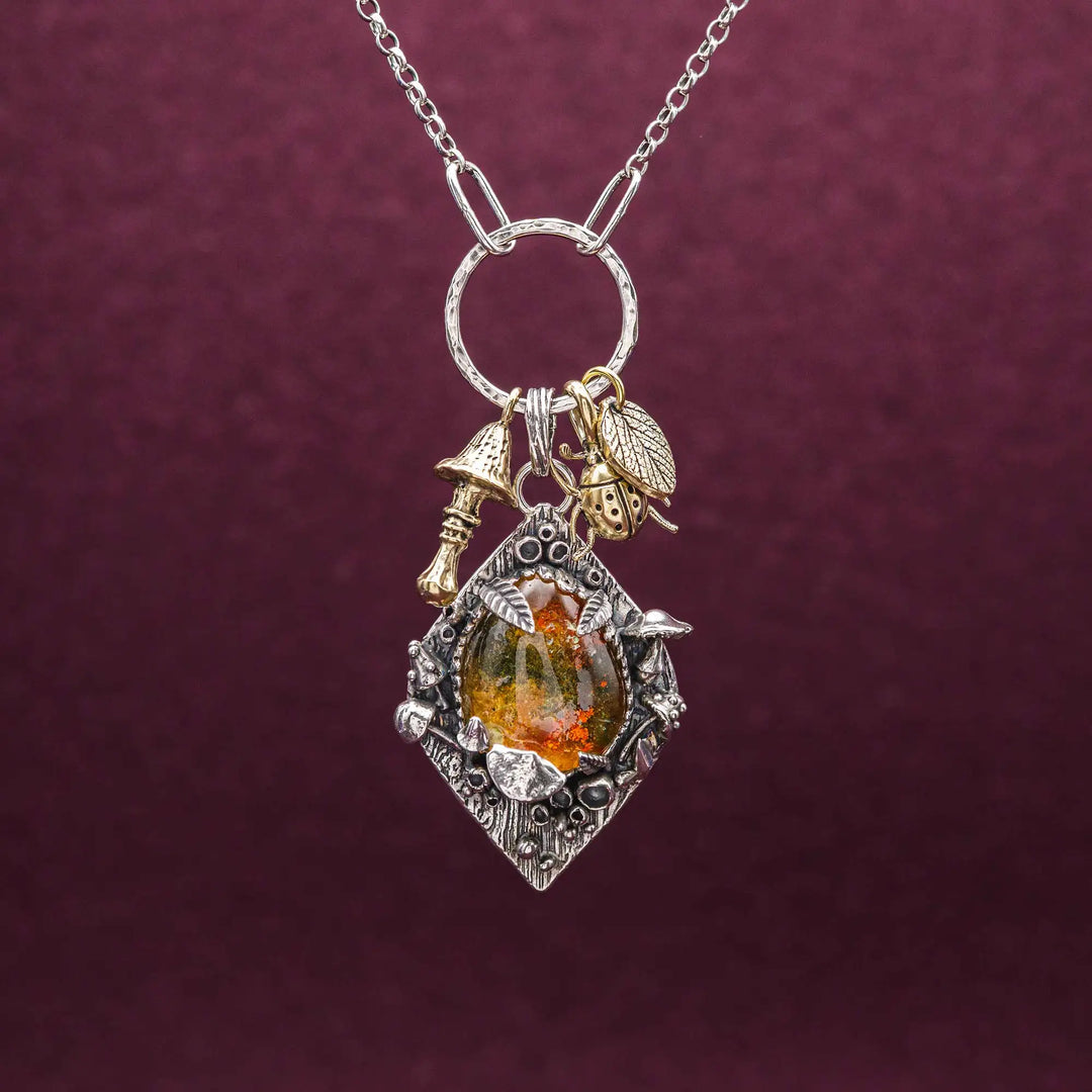Statement Kette groß aus Silber 925. In den Anhänger eingefasst ist ein tropfenförmiger Lodolith, auch Gartenquarz genannt. Der Stein sieht aus wie eingeschlossenes grünes, gelbes und orangefarbenes Moos und ist umrandet von Pilzen, Blättern und Knospen. Ein echtes, handgefertigtes Statement Piece an Naturschmuck.
An dem Anhänger hängen außerdem 3 kleine Charms aus Messing: Ein Marienkäfer, ein Pilz und ein Blatt.