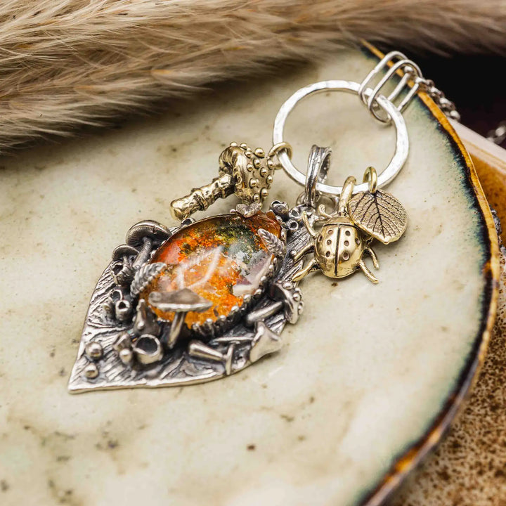 Statement Kette groß aus Silber 925. In den Anhänger eingefasst ist ein tropfenförmiger Lodolith, auch Gartenquarz genannt. Der Stein sieht aus wie eingeschlossenes grünes, gelbes und orangefarbenes Moos und ist umrandet von Pilzen, Blättern und Knospen. Ein echtes, handgefertigtes Statement Piece an Naturschmuck.
An dem Anhänger hängen außerdem 3 kleine Charms aus Messing: Ein Marienkäfer, ein Pilz und ein Blatt.